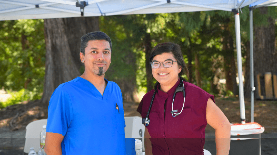 A man in blue scrubs smiles at the camera beside a woman in burgundy scrubs with a stethoscope around her neck, who is also smiling.